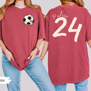 Op de afbeelding: Karmizijnrood T-shirt met een voetbalafbeelding en een hartje op de voorkant. De achterkant toont de naam "Pulisic" en het nummer "24" in cr&egrave;me. Het shirt heeft een comfortabele, oversized pasvorm. Het woord "Crimson" staat linksonder.