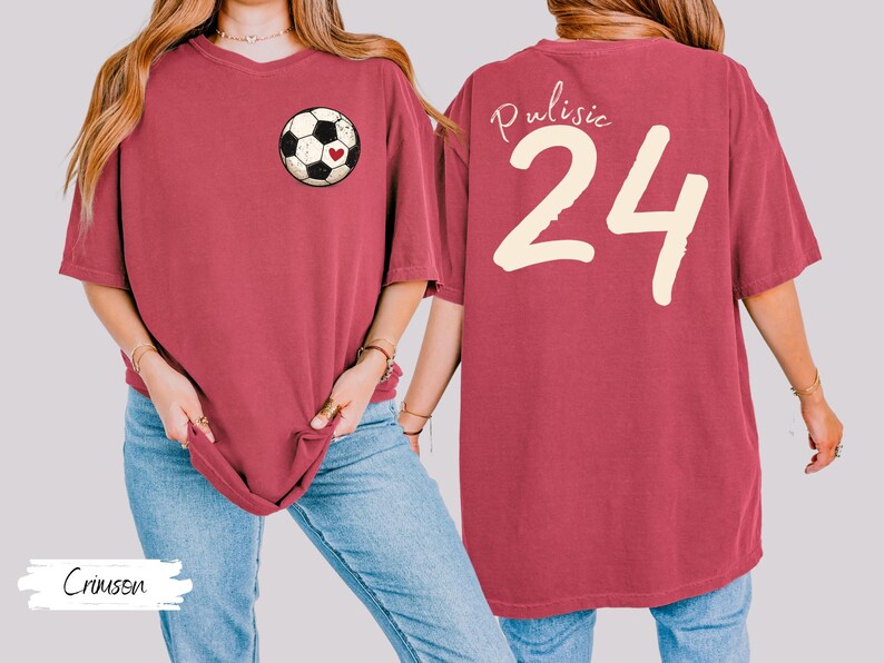 Op de afbeelding: Karmizijnrood T-shirt met een voetbalafbeelding en een hartje op de voorkant. De achterkant toont de naam "Pulisic" en het nummer "24" in cr&egrave;me. Het shirt heeft een comfortabele, oversized pasvorm. Het woord "Crimson" staat linksonder.