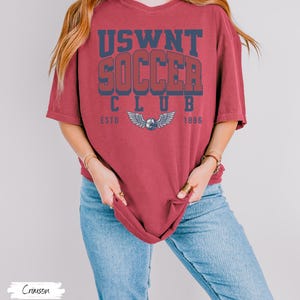 Puede incluir: Una camiseta roja carmes&iacute; con el texto "USWNT SOCCER CLUB ESTD 1886" en letras azul marino y rojo. La camiseta presenta un gr&aacute;fico de un bal&oacute;n de f&uacute;tbol con alas.