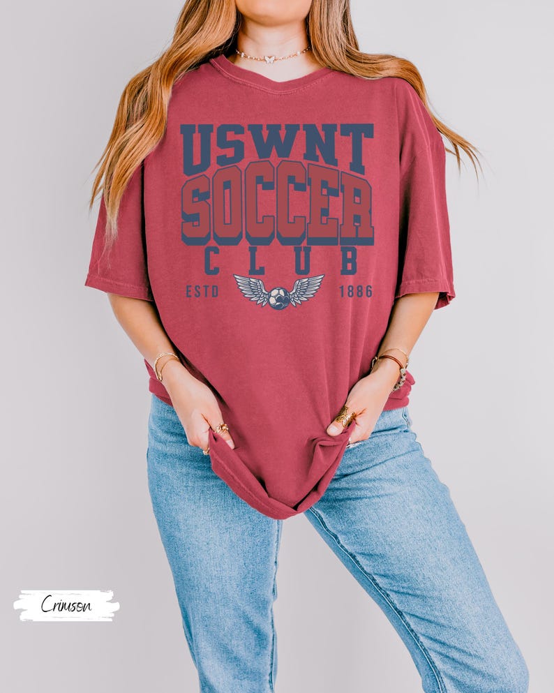 Puede incluir: Una camiseta roja carmes&iacute; con el texto "USWNT SOCCER CLUB ESTD 1886" en letras azul marino y rojo. La camiseta presenta un gr&aacute;fico de un bal&oacute;n de f&uacute;tbol con alas.