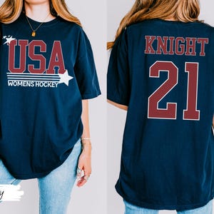 May include: Navy blue t-shirt with "USA WOMENS HOCKEY" in maroon and white. The front design includes stars. The back of the shirt displays "KNIGHT 21" in maroon. A casual, sporty tee.