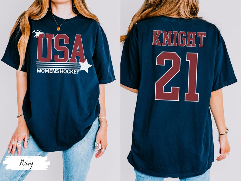 May include: Navy blue t-shirt with "USA WOMENS HOCKEY" in maroon and white. The front design includes stars. The back of the shirt displays "KNIGHT 21" in maroon. A casual, sporty tee.