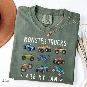 May include: An olive green t-shirt featuring the text "MONSTER TRUCKS ARE MY JAM" and illustrations of monster trucks. The shirt has a crew neck and short sleeves. A gold necklace and a brown hat are also in the image.