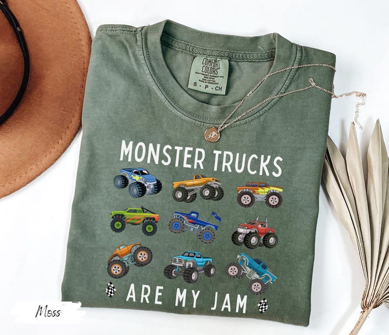 May include: An olive green t-shirt featuring the text "MONSTER TRUCKS ARE MY JAM" and illustrations of monster trucks. The shirt has a crew neck and short sleeves. A gold necklace and a brown hat are also in the image.