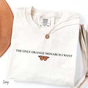 K&ouml;nnte beinhalten: Cremefarbenes T-Shirt mit dem Text "THE ONLY ORANGE MONARCH I WANT" und einer Schmetterlingsgrafik. Eine goldene Halskette mit Anh&auml;nger liegt auf dem Shirt. Ein brauner Hut und getrocknete Palmbl&auml;tter sind im Hintergrund.