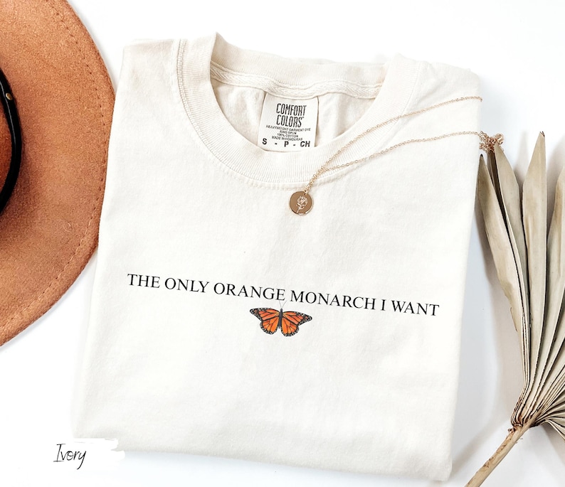 K&ouml;nnte beinhalten: Cremefarbenes T-Shirt mit dem Text "THE ONLY ORANGE MONARCH I WANT" und einer Schmetterlingsgrafik. Eine goldene Halskette mit Anh&auml;nger liegt auf dem Shirt. Ein brauner Hut und getrocknete Palmbl&auml;tter sind im Hintergrund.