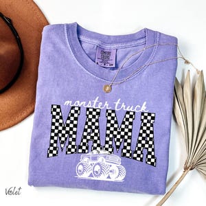 May include: A violet Comfort Colors t-shirt with the words "monster truck MAMA" in white and black checkered letters. A white monster truck graphic is below the text. A gold necklace and a brown hat are in the background.