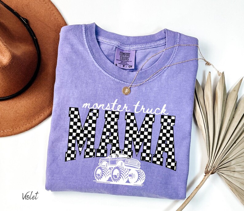 May include: A violet Comfort Colors t-shirt with the words "monster truck MAMA" in white and black checkered letters. A white monster truck graphic is below the text. A gold necklace and a brown hat are in the background.