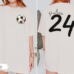 Op de afbeelding: Ivoor kleurig T-shirt met een voetbalafbeelding en een rood hart. De achterkant van het shirt toont de naam "Pulisic" boven het nummer "24" in zwart. Het shirt heeft een casual, oversized pasvorm.