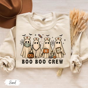 May include: A sand-colored sweatshirt featuring a graphic of four ghost characters dressed as medical professionals. The ghosts are wearing nurse hats and carrying medical tools. The text "BOO BOO CREW" is printed below the ghosts. A Halloween-themed design.
