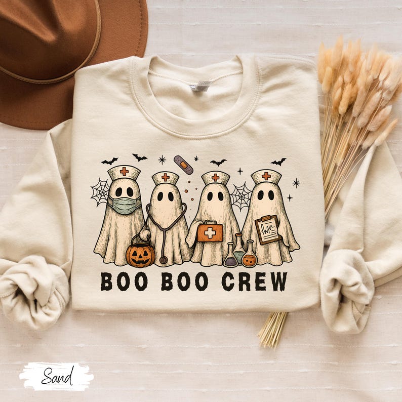 May include: A sand-colored sweatshirt featuring a graphic of four ghost characters dressed as medical professionals. The ghosts are wearing nurse hats and carrying medical tools. The text "BOO BOO CREW" is printed below the ghosts. A Halloween-themed design.