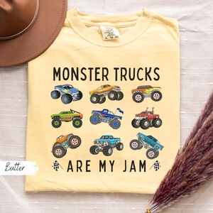 May include: A pale yellow t-shirt featuring the phrase "MONSTER TRUCKS ARE MY JAM" and images of various monster trucks. The tee has a round neck and short sleeves.