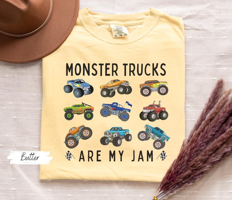 May include: A pale yellow t-shirt featuring the phrase "MONSTER TRUCKS ARE MY JAM" and images of various monster trucks. The tee has a round neck and short sleeves.
