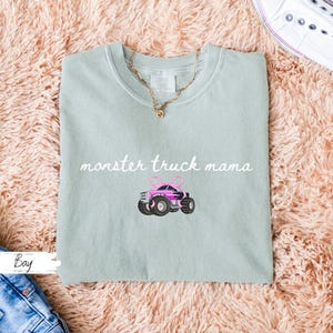 Puede incluir: Camiseta verde claro con la frase "monster truck mama" en cursiva. Debajo, un cami&oacute;n monstruo rosa bordado con ruedas negras y un lazo rosa. Se ve un collar dorado.