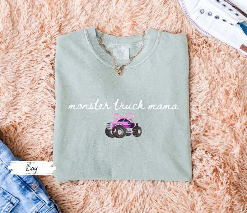 Puede incluir: Camiseta verde claro con la frase "monster truck mama" en cursiva. Debajo, un cami&oacute;n monstruo rosa bordado con ruedas negras y un lazo rosa. Se ve un collar dorado.