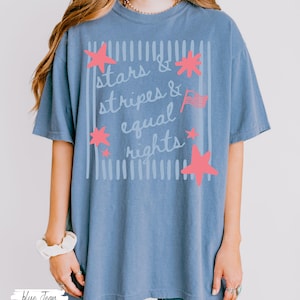 Puede incluir: Una camiseta azul vaquero con las palabras "stars & stripes & equal rights" en un dise&ntilde;o blanco y rosa. El dise&ntilde;o incluye estrellas rosas y una peque&ntilde;a bandera americana. La camiseta tiene un corte holgado y c&oacute;modo.