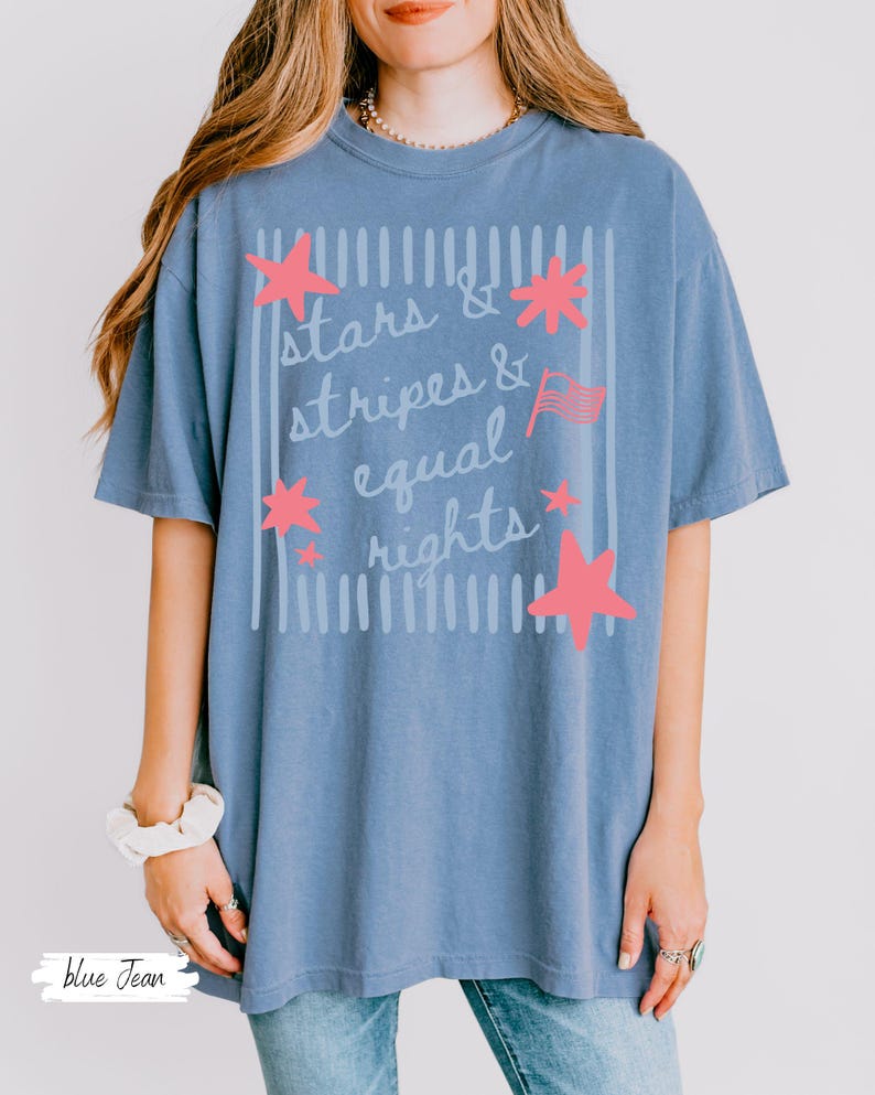 Puede incluir: Una camiseta azul vaquero con las palabras "stars & stripes & equal rights" en un dise&ntilde;o blanco y rosa. El dise&ntilde;o incluye estrellas rosas y una peque&ntilde;a bandera americana. La camiseta tiene un corte holgado y c&oacute;modo.