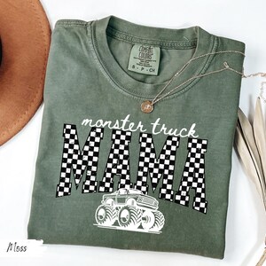 May include: A moss green t-shirt with the words "monster truck MAMA" in white, with a checkered pattern and a monster truck graphic. The shirt has a crew neck and short sleeves. A gold necklace is draped across the shirt.