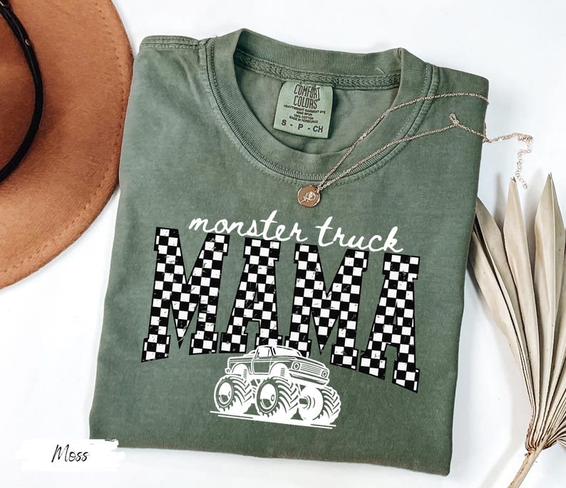 May include: A moss green t-shirt with the words "monster truck MAMA" in white, with a checkered pattern and a monster truck graphic. The shirt has a crew neck and short sleeves. A gold necklace is draped across the shirt.