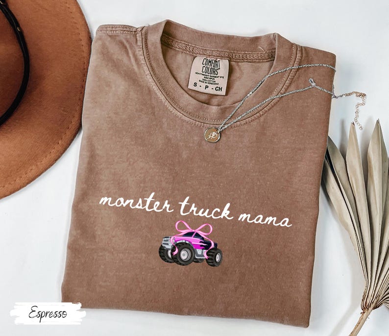 Puede incluir: Camiseta color espresso con las palabras "monster truck mama" en escritura blanca y un gr&aacute;fico de monster truck rosa y negro. Un collar plateado con un colgante est&aacute; sobre la camiseta.