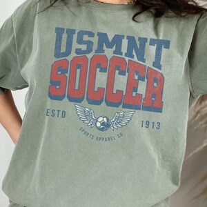 May include: Olive green t-shirt with the text "USMNT SOCCER" in blue and red, featuring a soccer ball with wings and the text "SPORTS APPAREL CO" and "ESTD 1913".