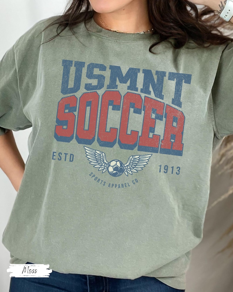 May include: Olive green t-shirt with the text "USMNT SOCCER" in blue and red, featuring a soccer ball with wings and the text "SPORTS APPAREL CO" and "ESTD 1913".