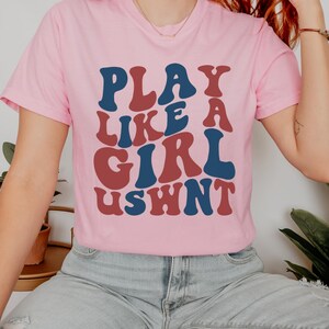 May include: A pink t-shirt with the text "PLAY LIKE A GIRL USWNT" in a retro font style. The text is in red and blue.