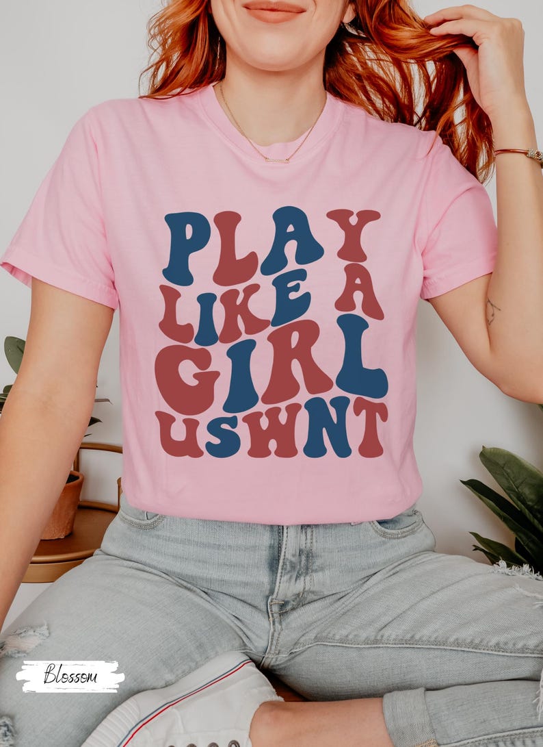 May include: A pink t-shirt with the text "PLAY LIKE A GIRL USWNT" in a retro font style. The text is in red and blue.