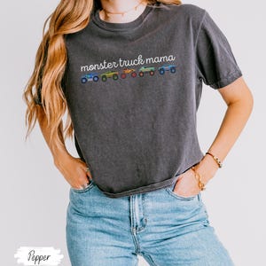 May include: Dark gray t-shirt with the text "monster truck mama" in white script above a row of colorful monster truck illustrations. The shirt is paired with light blue jeans.