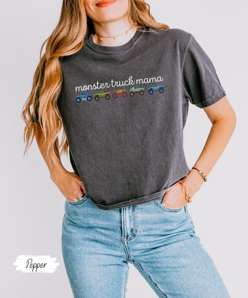 May include: Dark gray t-shirt with the text "monster truck mama" in white script above a row of colorful monster truck illustrations. The shirt is paired with light blue jeans.