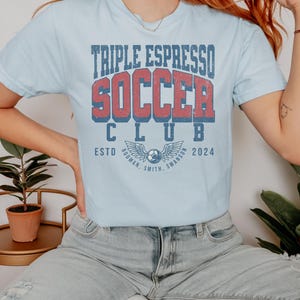 Op de afbeelding: Een lichtblauw T-shirt met de tekst "Triple Espresso Soccer Club" in rood en blauwe letters. De tekst "Estd 2024" staat onder de clubnaam. Het shirt heeft een vintage-stijl afbeelding van een voetbal met vleugels.