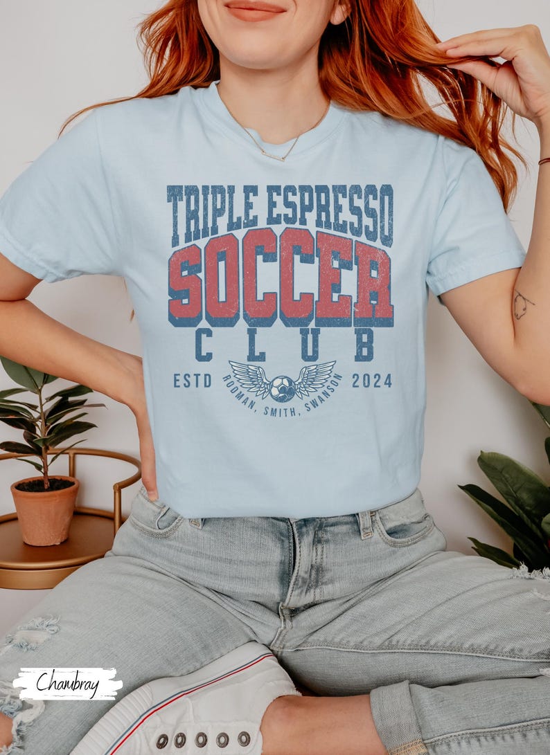Op de afbeelding: Een lichtblauw T-shirt met de tekst "Triple Espresso Soccer Club" in rood en blauwe letters. De tekst "Estd 2024" staat onder de clubnaam. Het shirt heeft een vintage-stijl afbeelding van een voetbal met vleugels.