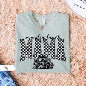 May include: A light green t-shirt with the words "monster truck MAMA" in black, checkered letters. A black and white monster truck graphic is below the text. A gold necklace is visible at the neckline.