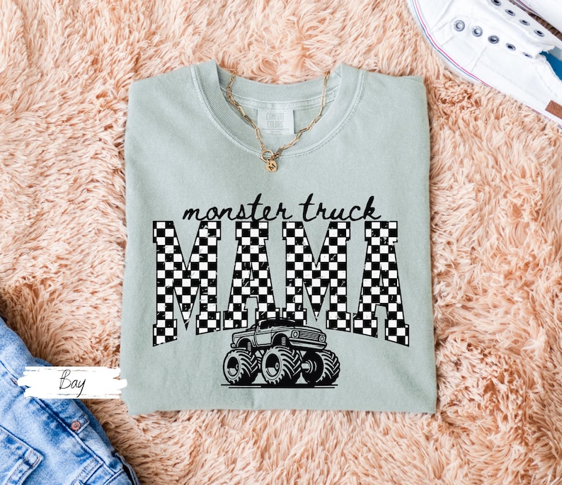 May include: A light green t-shirt with the words "monster truck MAMA" in black, checkered letters. A black and white monster truck graphic is below the text. A gold necklace is visible at the neckline.