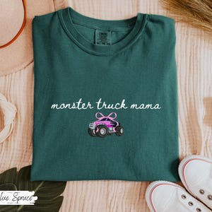 Puede incluir: Camiseta verde oscuro con la frase "monster truck mama" en cursiva y un gr&aacute;fico de un cami&oacute;n monstruo rosa. La camiseta est&aacute; doblada y expuesta sobre una superficie color crema.
