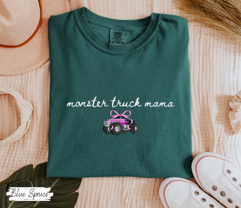 Puede incluir: Camiseta verde oscuro con la frase "monster truck mama" en cursiva y un gr&aacute;fico de un cami&oacute;n monstruo rosa. La camiseta est&aacute; doblada y expuesta sobre una superficie color crema.