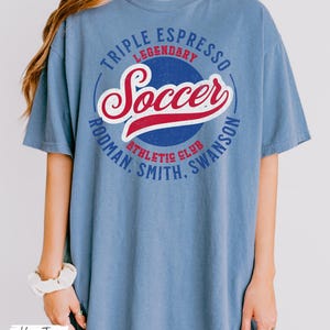 Puede incluir: Una camiseta de gran tama&ntilde;o de color azul vaquero con un dise&ntilde;o gr&aacute;fico de estilo vintage que incluye el texto "Triple Espresso Legendary Soccer Rodman. Smith. Swanson Athletic Club".
