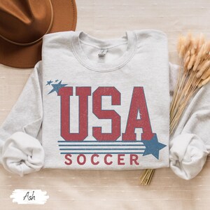 May include: A gray sweatshirt with a vintage-style graphic of "USA SOCCER" in red and blue with stars.