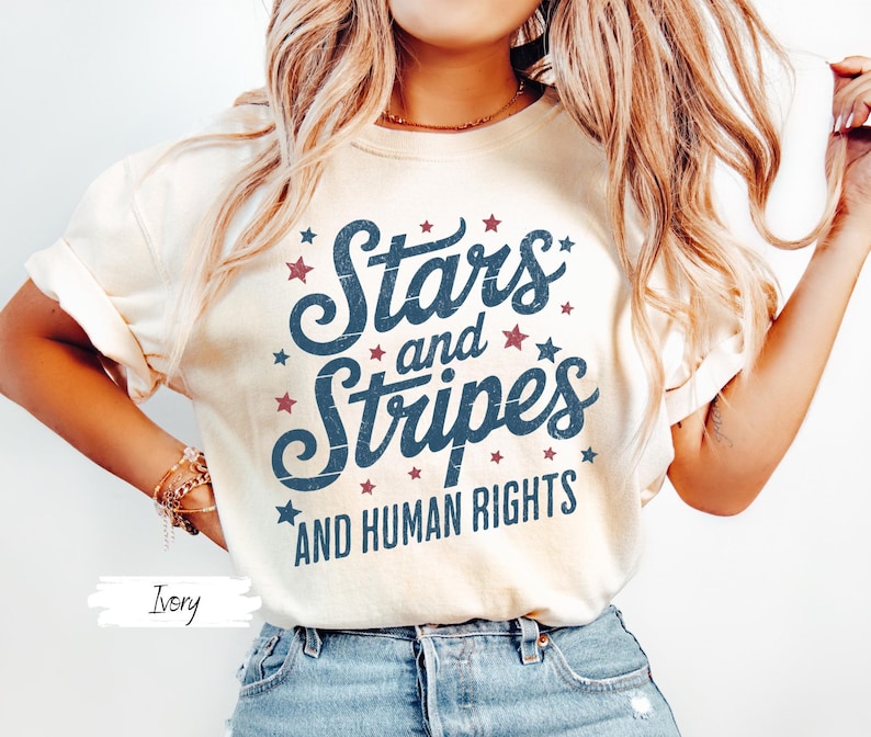 Puede incluir: Camiseta color marfil con el texto "Stars and Stripes AND HUMAN RIGHTS" en azul marino y estrellas rojas. Una camiseta de estilo casual, adecuada para el uso diario.