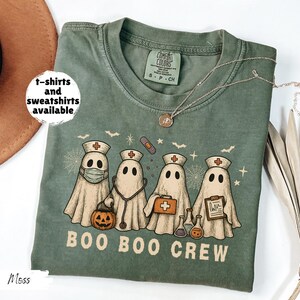 May include: Moss green t-shirt with a graphic of four ghost characters dressed as nurses, with a pumpkin, stethoscope, and medical supplies. The text "BOO BOO CREW" is below the ghosts. The shirt has a crew neck and short sleeves. "t-shirts and sweatshirts available" is written on the left.