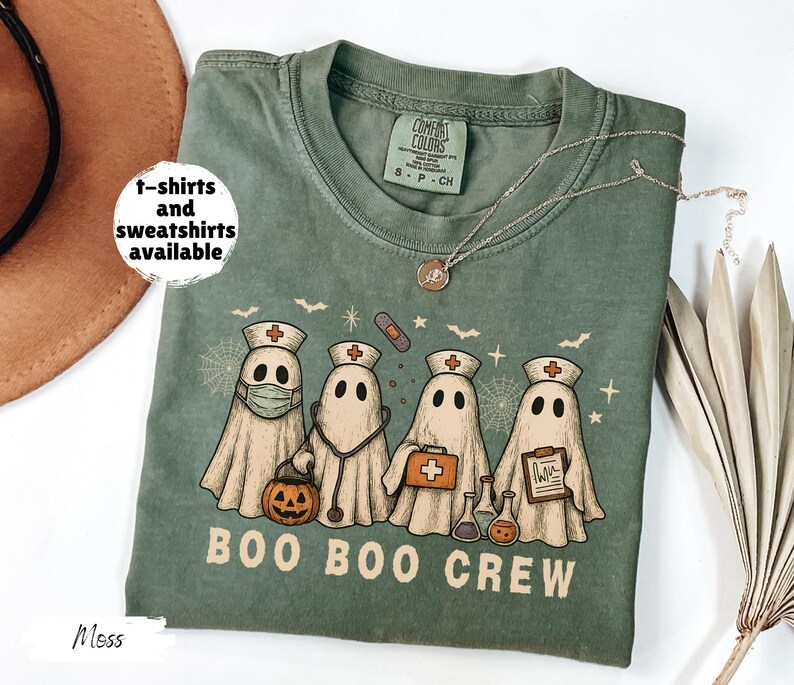 May include: Moss green t-shirt with a graphic of four ghost characters dressed as nurses, with a pumpkin, stethoscope, and medical supplies. The text "BOO BOO CREW" is below the ghosts. The shirt has a crew neck and short sleeves. "t-shirts and sweatshirts available" is written on the left.