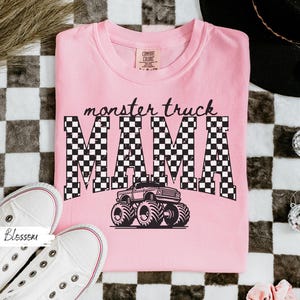 Op de afbeelding: Een lichtroze t-shirt met de tekst "monster truck MAMA" in een zwart-wit geblokt patroon en een monster truck afbeelding. Witte sneakers liggen ernaast.