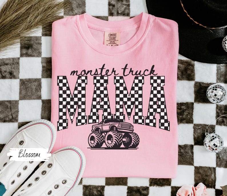 Op de afbeelding: Een lichtroze t-shirt met de tekst "monster truck MAMA" in een zwart-wit geblokt patroon en een monster truck afbeelding. Witte sneakers liggen ernaast.