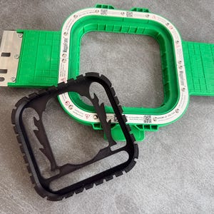 May include: A green and white embroidery hoop with a black frame. The hoop has a square shape and is made of plastic. The black frame is designed to fit inside the hoop. The hoop is designed for use with embroidery machines.