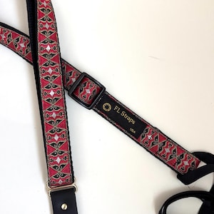 May include: A red and black guitar strap with a repeating geometric pattern. The strap has black leather ends and a black adjustable buckle. The text "FL Straps USA" is visible on the strap.