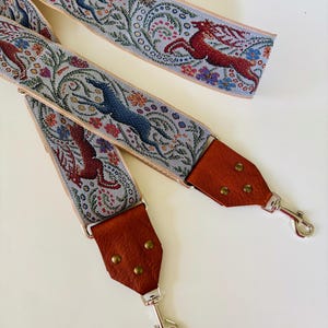 May include: A decorative bag strap with a woven design featuring leaping deer in red and blue, surrounded by floral patterns. The strap has brown leather ends with metal clasps for attachment.