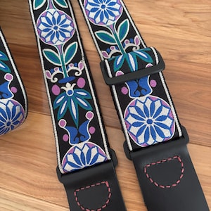 Könnte beinhalten: Schwarzer Gitarrengurt mit einem floralen Design in Blau, Lila und Grün. Der Gurt hat ein sich wiederholendes Muster aus stilisierten Blumen und Blättern auf schwarzem Hintergrund. Die Enden sind aus schwarzem Leder mit rosa Nähten und verstellbaren schwarzen Kunststoffschnallen.