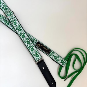 May include: A guitar strap with a white background. The strap is white with a green floral pattern. The strap has black leather ends and silver metal hardware. The text "FL Straps USA" is on a black tag.
