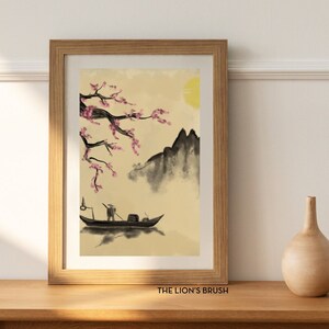 Puede incluir: Una impresión enmarcada de una pintura japonesa tradicional que representa un barco con una persona en él, una cordillera y un cerezo en flor con flores rosas. La impresión está sobre un fondo beige y tiene un sol amarillo claro en la esquina superior derecha. El marco es de madera marrón y la impresión está titulada "THE LION'S BRUSH".