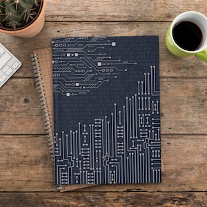 May include: A black notebook with a white circuit board design and binary code. The notebook is closed and has a spiral binding. The notebook is on a wooden table with a white keyboard and a green coffee mug.